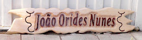 Placa longa de cedro branco com bordas orgânicas e o nome "João Orides Nunes" entalhado em alto relevo, com logotipos da fazenda em baixo relevo nas laterais