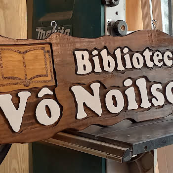 Placa de madeira maciça com o texto "Biblioteca Vô Nilson" em letras brancas em relevo e um livro aberto pirografado na lateral esquerda