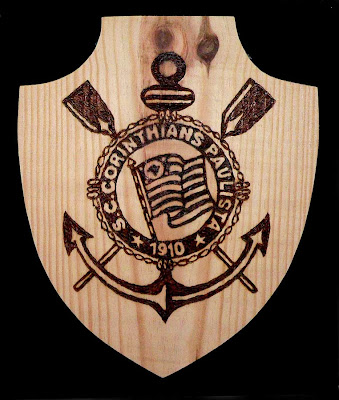 Escudo de madeira clara com o brasão do Corinthians, com âncora, remos e a bandeira, gravado em pirografia