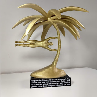 Troféu de resina dourada com um coqueiro e a figura de um atleta em movimento, sobre base preta com frase gravada