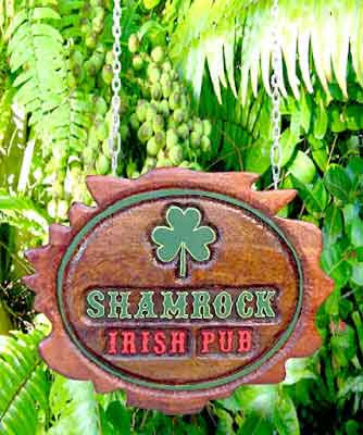 Placa oval suspensa em cedro escuro com trevo e as palavras "Shamrock Irish Pub" entalhadas em altíssimo alto relevo e pintadas em verde e vermelho, pendurada por correntes entre folhagens