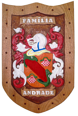 Brasão da Família Andrade entalhado em madeira, com escudo laranja, elmo branco, folhagens vermelhas e faixas com os nomes