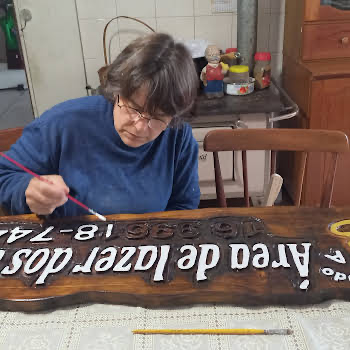 Artesã pintando à mão, com pincel fino, as letras de uma placa de madeira entalhada com o texto "Área de lazer" sobre a mesa do ateliê