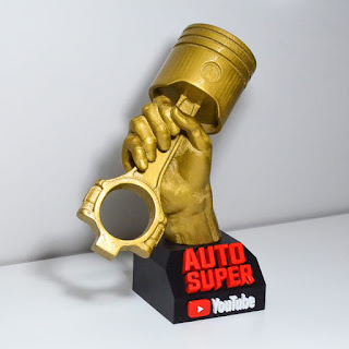 Troféu de resina dourada em forma de mão segurando um pistão de motor, sobre base preta com a marca Auto Super