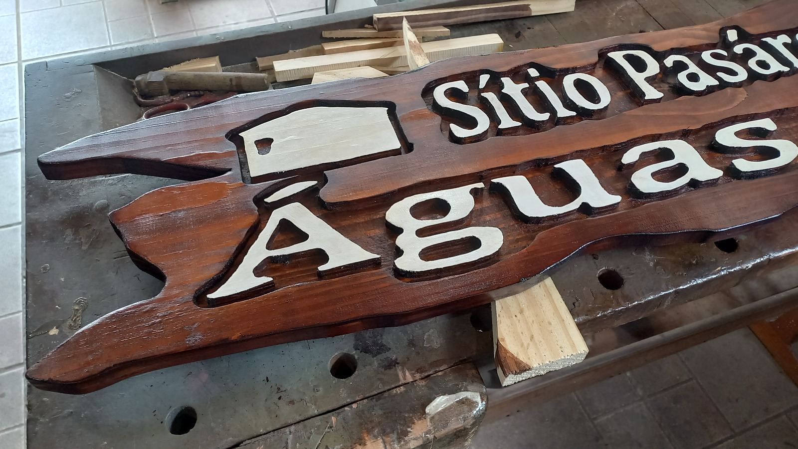 Placa de madeira maciça tonalizada em nogueira com letras brancas em alto relevo escritas Sitio Pasargada das Aguas, sobre bancada de marcenaria