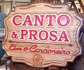 Placa de madeira pinus com o texto "Canto & Prosa, Com o Cancioneiro" em alto relevo pintado em vermelho e roda de carreta entalhada