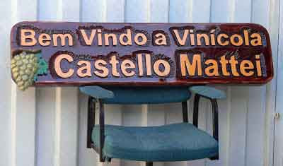 Placa horizontal em madeira maciça com as letras "Bem Vindo a Vinícola Castello Mattei" em alto relevo e um cacho de uvas esculpido em super alto relevo, com acabamento espelhado