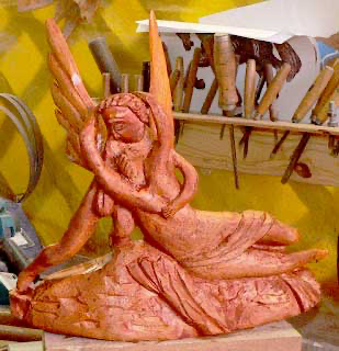 Escultura de anjo com asas e vestes esculpidas em madeira, sobre a bancada da oficina com formões ao fundo