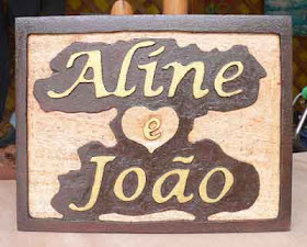 Placa retangular de pinus com moldura escura e os nomes "Aline e João" entalhados em alto relevo com um coração no centro
