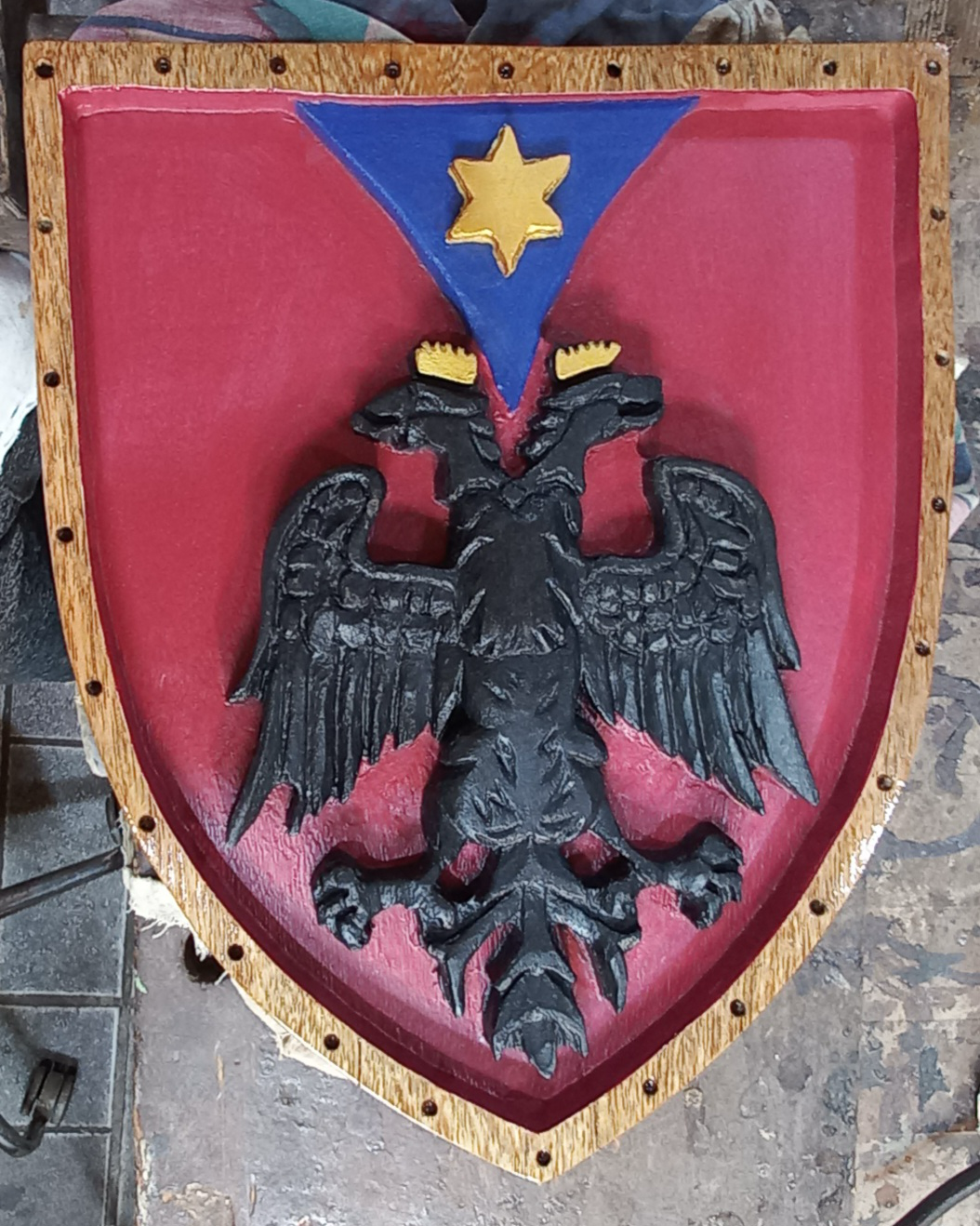 Brasão de família entalhado em madeira maciça em formato de escudo, com águia bicéfala preta em alto relevo, campo vermelho, cunha azul e estrela dourada de seis pontas