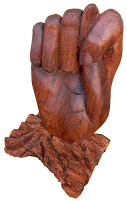 Escultura de uma mão fechada em madeira avermelhada, apoiada em base de madeira bruta