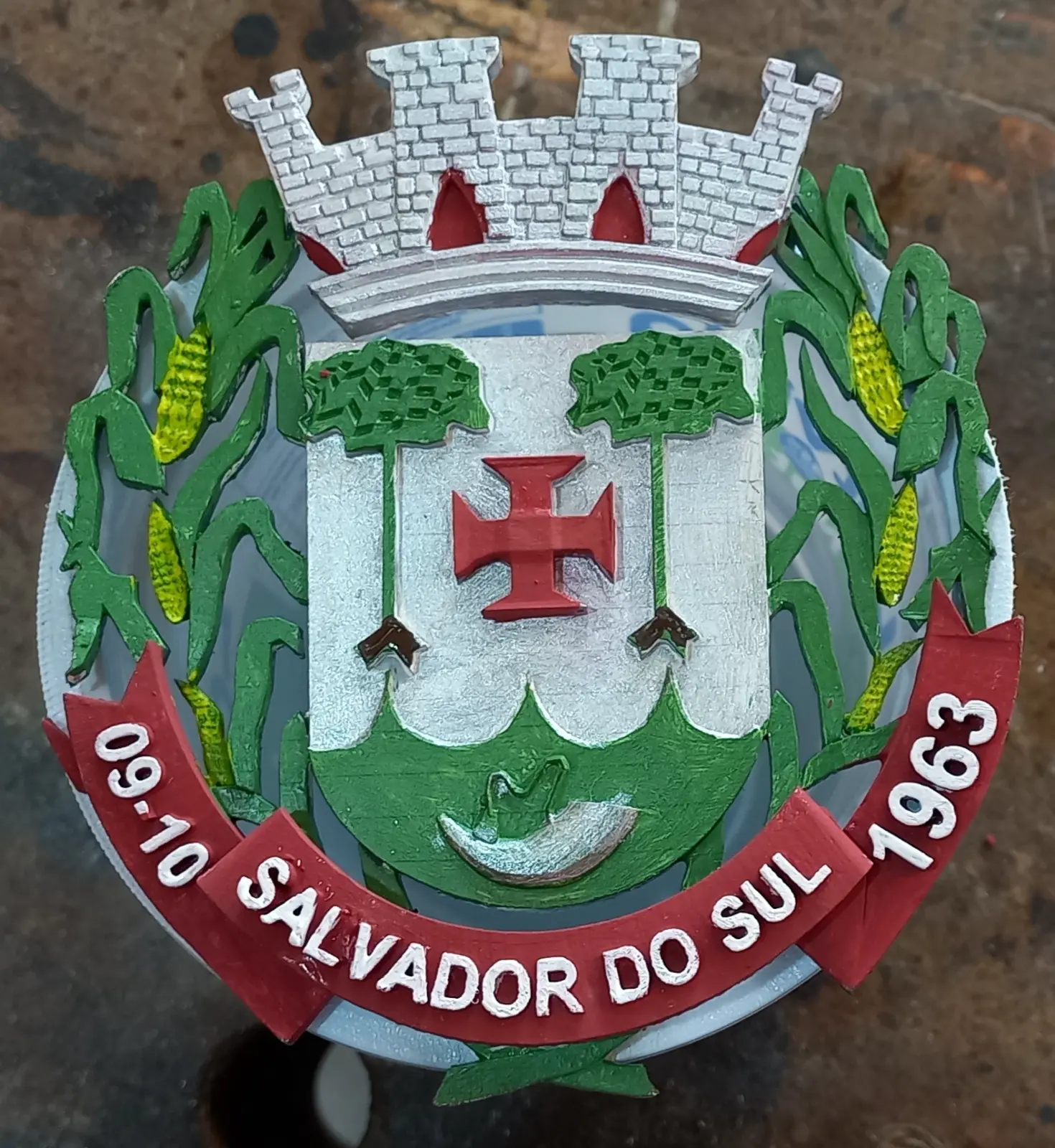 Brasão do município de Salvador do Sul em relevo, com coroa mural, cruz vermelha, espigas de milho e faixa com a data 09-10-1963, pintado à mão