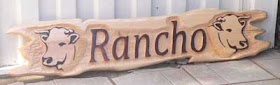 Placa longa de cedro branco com a palavra "Rancho" em alto relevo e duas cabeças de boi entalhadas e escurecidas com pirografia