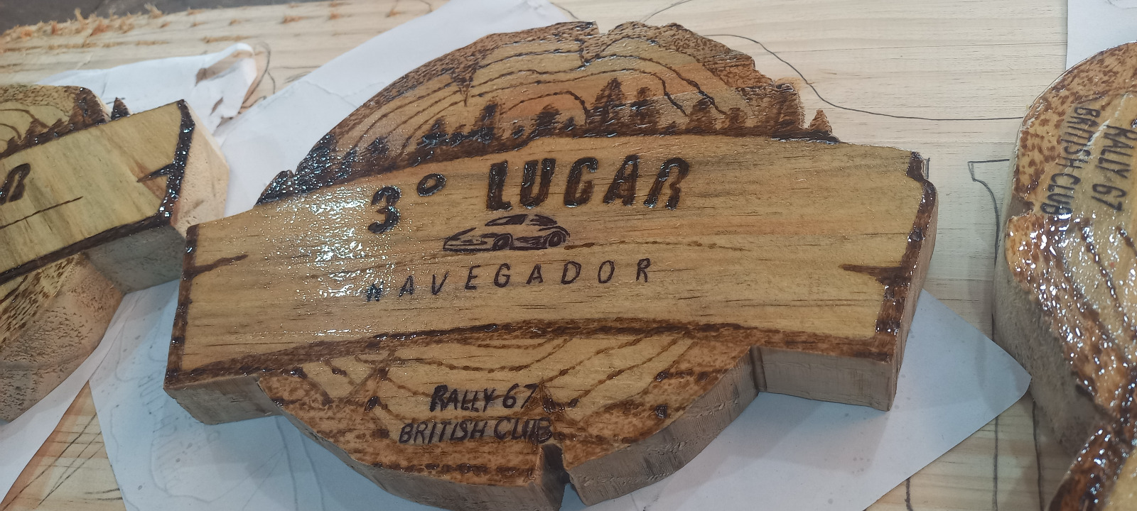 Detalhe aproximado do troféu "3º Lugar Navegador" do Rally 67 British Club mostrando a pirografia da paisagem, o carro e o brilho do verniz marítimo