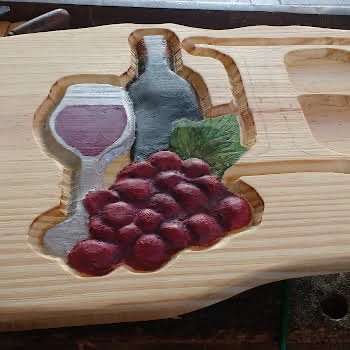 Detalhe de placa em madeira clara com entalhe em alto relevo de garrafa de vinho, taça e cacho de uvas pintado à mão