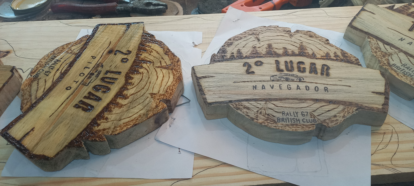 Troféus de 2º lugar piloto e navegador do Rally 67 British Club em madeira maciça pirografada, com paisagem de serra e carro de rally, sobre a bancada do ateliê