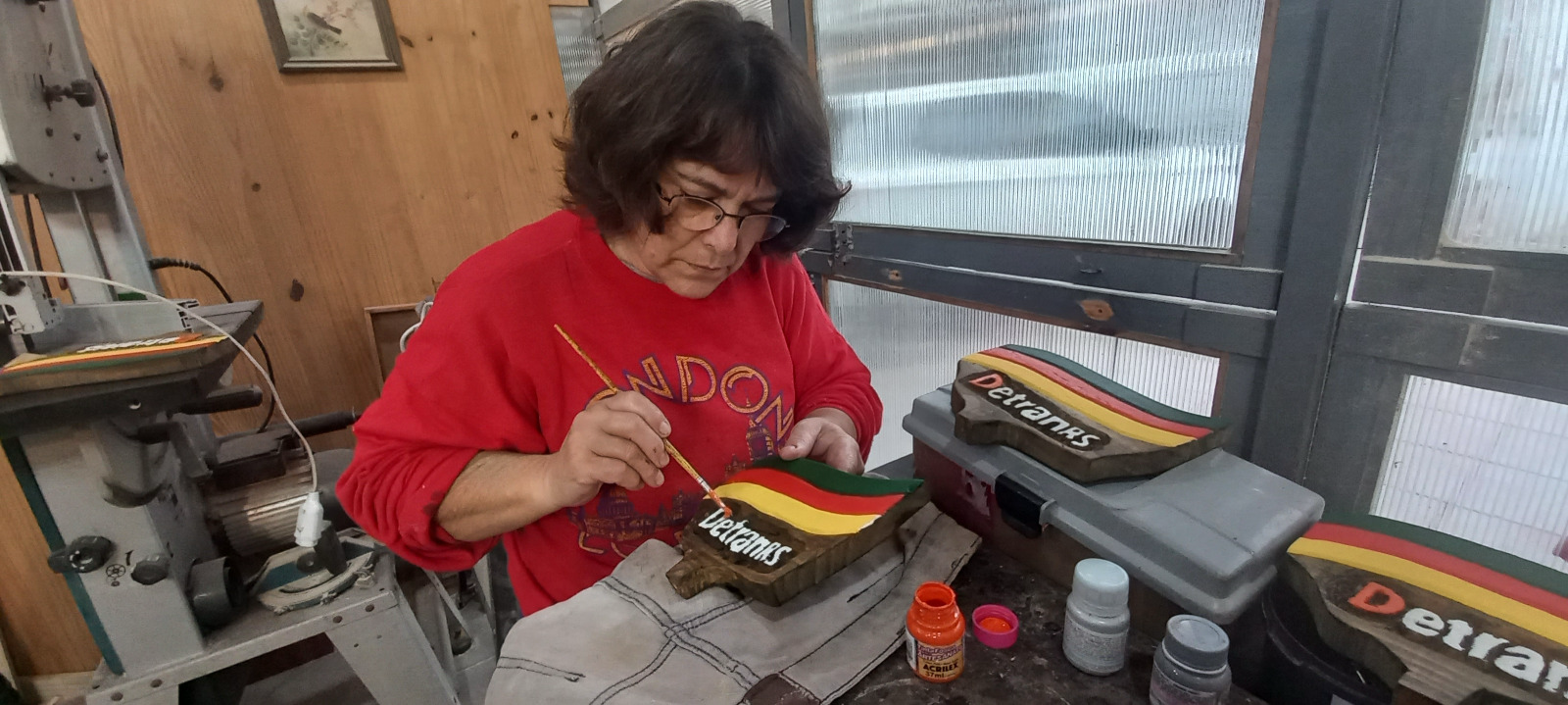 Artesã pintando à mão, com pincel fino, as letras de um troféu em madeira do DETRAN RS na bancada do ateliê De Moraes