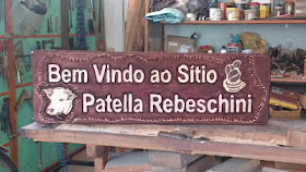 Placa em cedro branco escurecido com anilina, com o texto "Bem Vindo ao Sítio Patella Rebeschini" em letras de alto relevo e logotipo com cabeça de boi pirografada