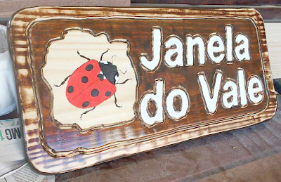 Placa de madeira pinus tonalizada com o nome "Janela do Vale" em letras brancas em alto relevo e uma joaninha pintada à mão em painel claro