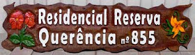 Placa horizontal de cedro com o texto "Residencial Reserva Querência nº 855" em alto relevo e flores pintadas em tinta acrílica