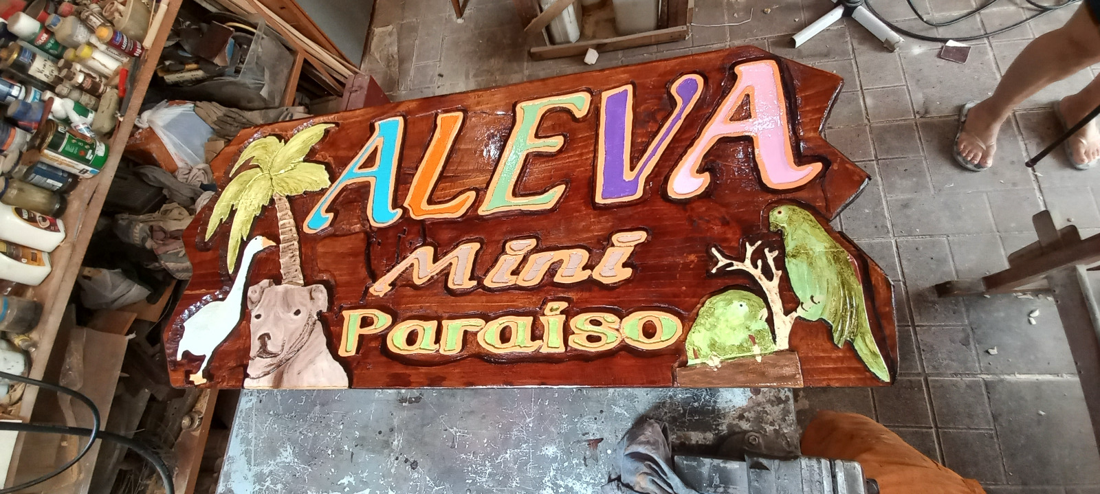 Painel de madeira entalhado com o texto "Aleva Mini Paraíso" em letras coloridas em alto relevo 3D, com coqueiro, ganso, cachorro e dois papagaios esculpidos e pintados
