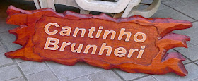 Placa de pinus com bordas orgânicas avermelhadas e o texto "Cantinho Brunheri" entalhado em alto e baixo relevo pintado de branco