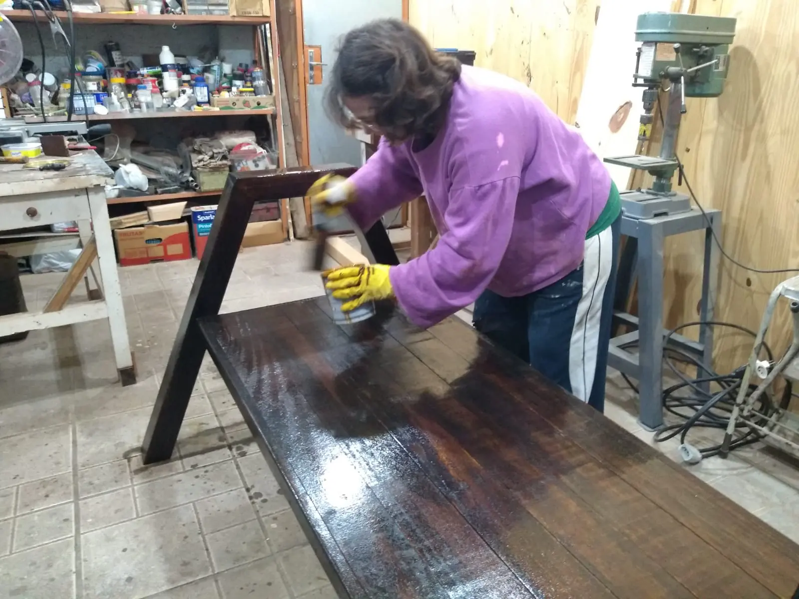 Artesã aplicando verniz à mão sobre o assento do banco rústico de madeira maciça dentro da marcenaria