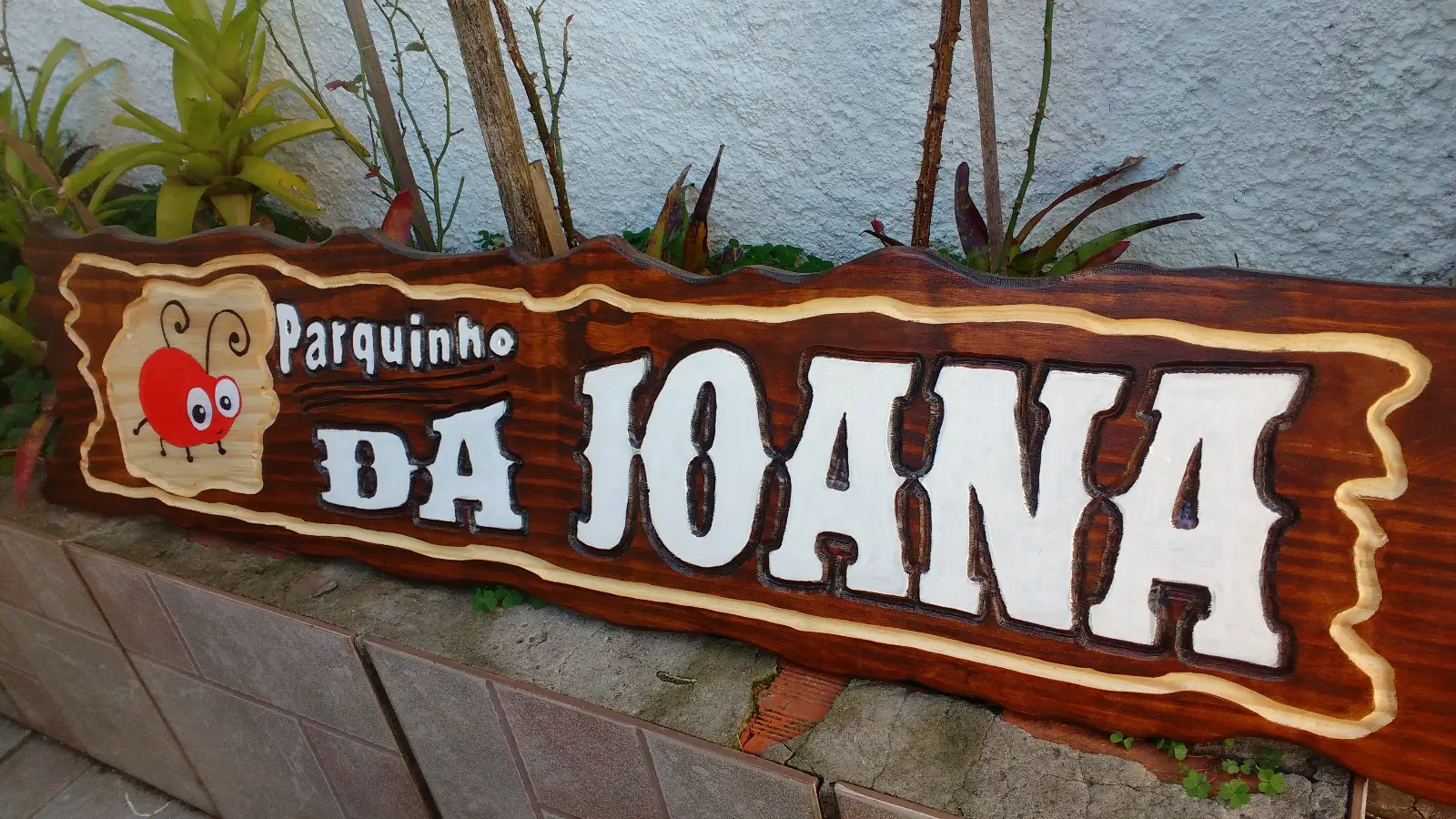 Placa de madeira tonalizada com letras brancas em alto relevo escritas "Parquinho da Joana" e desenho de uma joaninha vermelha pintada à mão