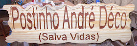 Placa horizontal de madeira pinus com os textos "Postinho André Déco" e "(Salva Vidas)" entalhados em duplo baixo relevo