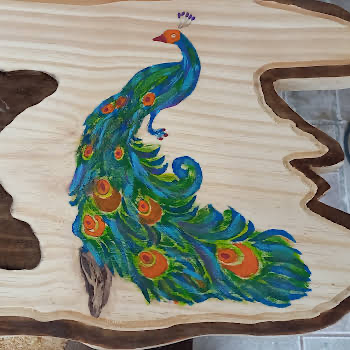 Pavão pintado à mão em tons de azul, verde e laranja sobre prancha de madeira clara com bordas naturais irregulares e casca escura
