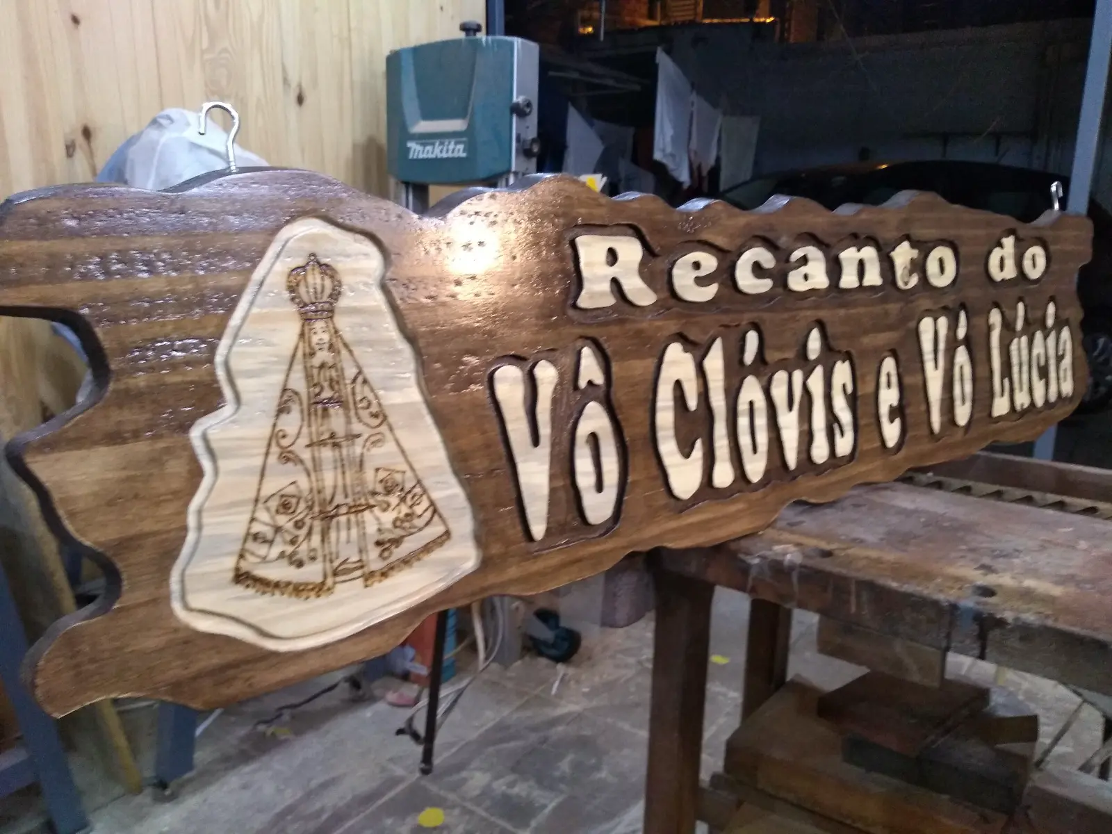 Placa de madeira tonalizada com bordas recortadas, letras claras em alto relevo escritas "Recanto do Vô Clóvis e Vó Lúcia" e imagem de Nossa Senhora Aparecida pirografada na madeira